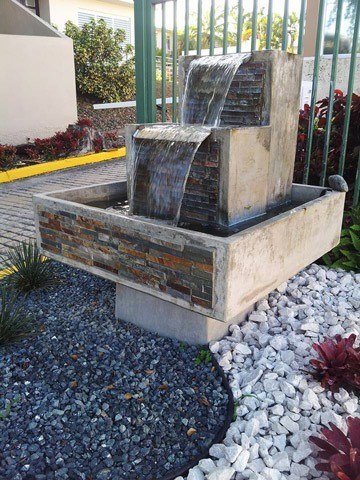fuente san francisco con piedras en los lados y montada en un jardin de piedras y plantas en la entrada de una urbanizacion.